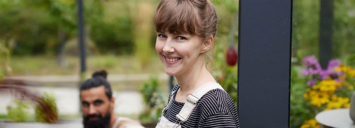 Happy woman wearing overalls is standing in the garden. A man sits in the background.