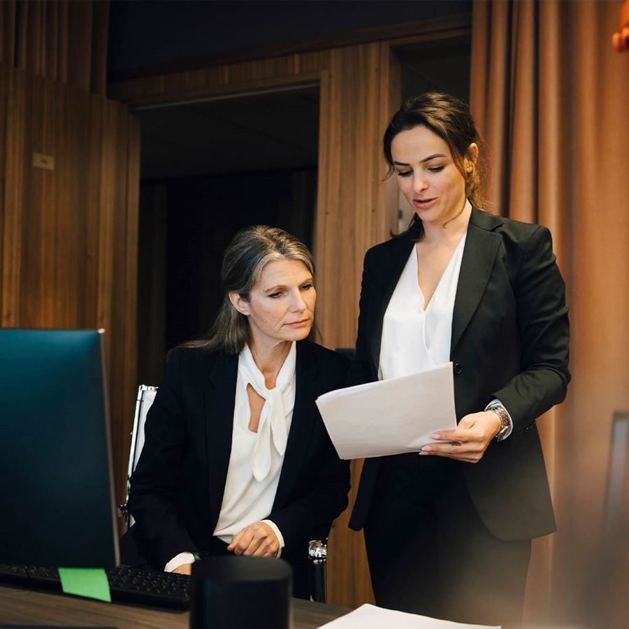 Two women in suits are discussing a contract. One is sitting down. 