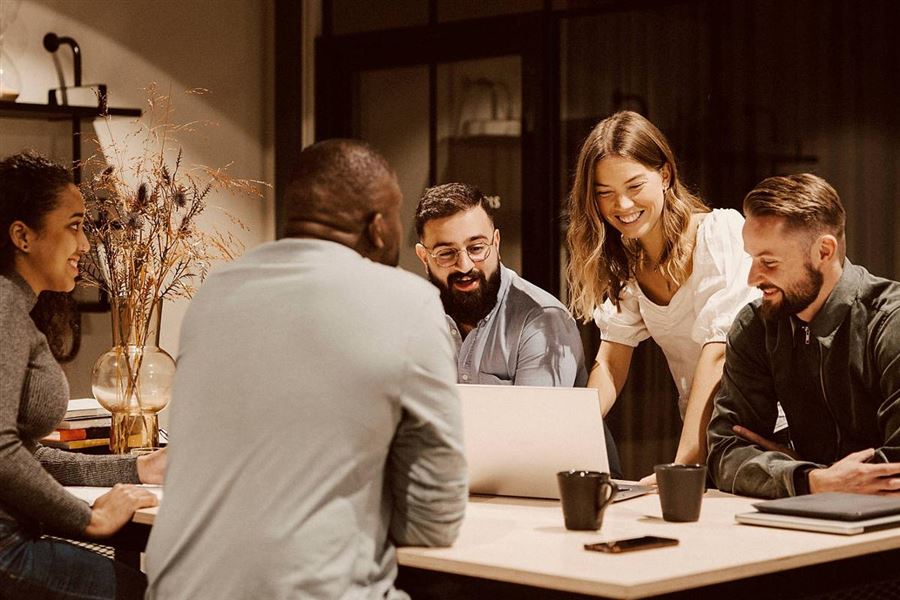 A group of colleagues are gathered around a table to discuss something. One person is showing his laptop screen to the others.