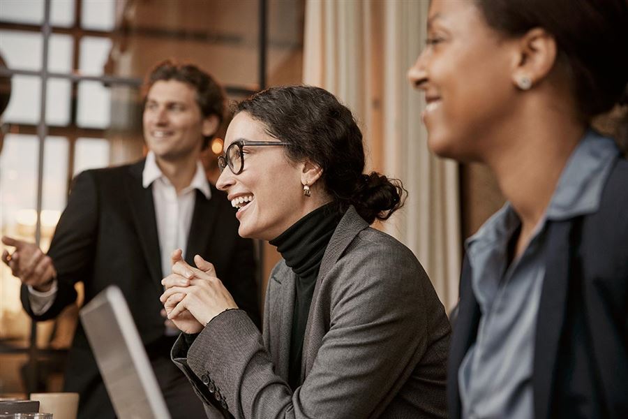 Three colleagues are sitting down at the office. They are laughing. 