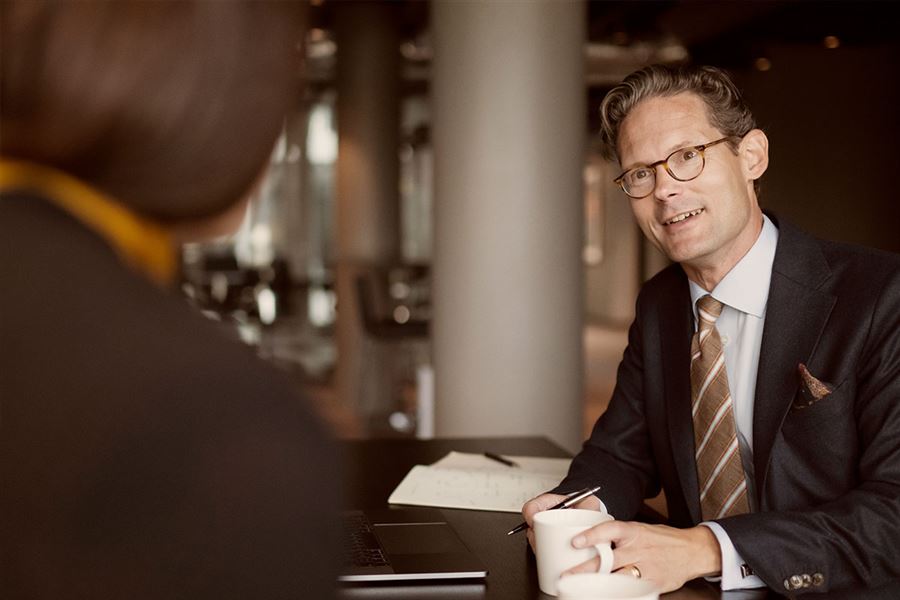 En rådgivare iklädd kostym har ett möte med en investerare. Han har en kopp kaffe i handen och ler.